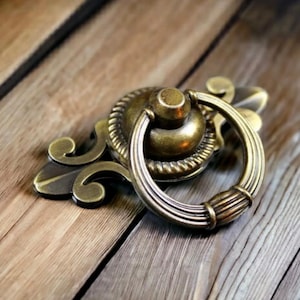 Puede incluir: Tirador de cajón de latón antiguo con un tirador de anillo y placa trasera ornamentada. El herraje presenta un acabado envejecido y detallado, diseñado para la restauración de muebles o uso decorativo. El tirador está sobre una superficie de madera.