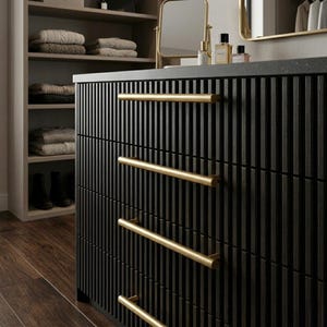 Könnte beinhalten: Eine schwarze Kommode mit vertikalen Rillen und goldfarbenen Stangengriffen. Die Kommode ist Teil eines Schlafzimmersets, mit einem Regal und einem Schminkspiegel im Hintergrund. Der Holzboden verleiht dem Raum Wärme.