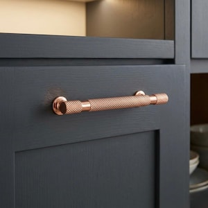 Puede incluir: Un tirador de armario de color cobre con un centro texturizado con forma de diamante y extremos lisos y redondeados. El herraje está montado en una puerta de armario gris oscuro. El tirador mide aproximadamente 15 cm de largo.