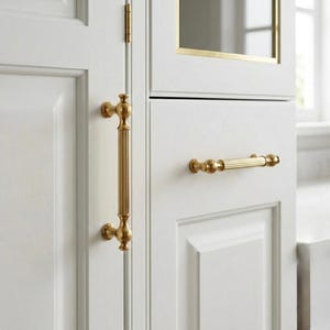 Puede incluir: Primer plano de puertas de armario blancas con herrajes dorados. Las puertas del armario tienen un diseño de paneles. Los herrajes incluyen un tirador largo y vertical y un tirador horizontal más corto. Los herrajes tienen un diseño decorativo y ornamentado.