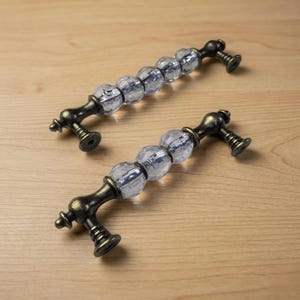 以下が含まれることがあります： アンティークスタイルのキャビネットプル2つ。各プルは、透明でファセット加工されたガラスビーズと、装飾的なダークブロンズ色の金属製ハードウェアを備えています。プルは家具やキャビネット用に設計されています。