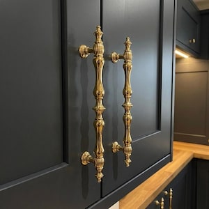 May include: Two ornate, gold-coloured cabinet pulls are mounted on a dark grey cabinet door. The pulls have a vertical, elongated design with decorative detailing. The cabinet door is part of a kitchen setup.