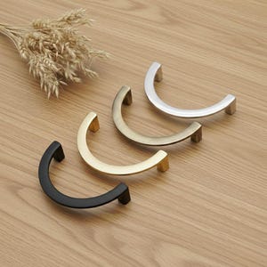 Peut inclure: Quatre poignées de meubles aux finitions métalliques variées : noir, doré, laiton et argent. Chaque poignée a une forme incurvée en demi-cercle avec une base rectangulaire. Elles sont présentées sur une surface en bois brun clair.