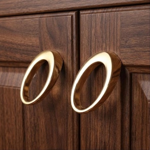 May include: Close-up of two wooden cabinet doors with oval-shaped, gold-colored handles. The doors are a dark brown color with visible wood grain. The handles are mounted on the doors and have a smooth, polished finish. The cabinet doors are part of a larger piece of furniture.