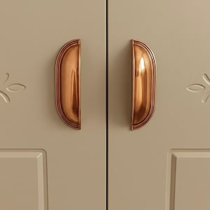 Könnte beinhalten: Nahaufnahme von zwei Schranktüren mit passenden, halbmondförmigen Griffen in Kupferfarbe. Die Griffe haben ein geriffeltes Design und sind an einem hellbeigen Schrank montiert. Florale Schnitzereien sind sichtbar.