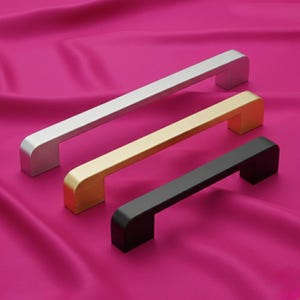 Könnte beinhalten: Drei Schrankgriffe in Silber, Gold und Schwarz, angeordnet auf einem rosa Stoff. Jeder Griff hat einen rechteckigen Stab mit abgerundeten Enden und einem quadratischen Sockel. Der silberne Griff befindet sich oben, der goldene in der Mitte und der schwarze unten.