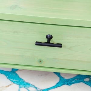 May include: Close-up of a black cabinet pull on a light green wooden drawer. The pull is a T-shaped design with a round knob and a horizontal bar. The drawer is part of a larger cabinet.