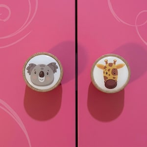 May include: Two round cabinet knobs with animal faces. One knob features a koala bear, the other a giraffe. The knobs have a white background with a wooden border. The cabinet doors are a bright pink color.