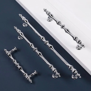 May include: A selection of polished chrome cabinet pulls and handles. The hardware has an ornate, vintage design with decorative detailing. The pulls vary in size and are arranged on a white and navy blue background.