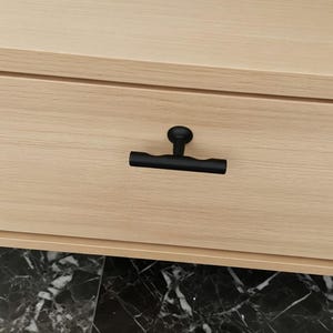 May include: Close-up of a black cabinet pull with a round knob and a horizontal bar. The pull is mounted on a light-colored wooden drawer. The drawer is part of a larger cabinet or dresser.