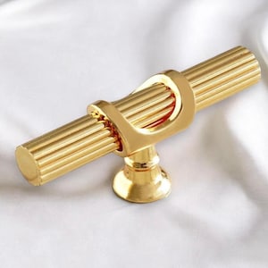 Könnte beinhalten: Ein goldfarbener Schrankgriff mit zylindrischem, geriffeltem Griff und dekorativem Ringdetail. Der Griff ist ca. 10 cm lang. Die Hardware ist aus Metall und für Möbel konzipiert.