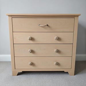 May include: A light wood dresser with four drawers. The top drawer has a rope handle, while the other three drawers have rope knot knobs. The dresser is made of light-colored wood and has a simple, modern design.