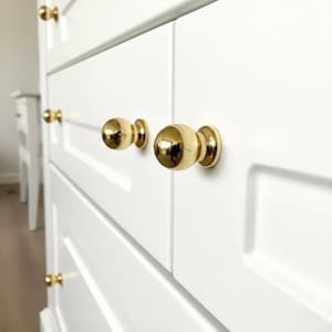 Könnte beinhalten: Nahaufnahme eines weißen Schranks mit goldenen, kugelförmigen Knöpfen. Die Knöpfe sind gleichmäßig angeordnet und reflektieren das Licht. Die Schranktüren haben ein eingelassenes Paneeldesign, das eine saubere, moderne Ästhetik schafft.