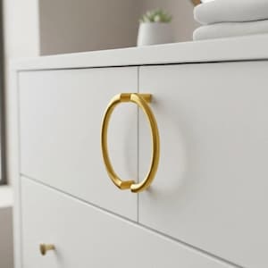 Puede incluir: Primer plano de una cómoda blanca con un tirador de anillo dorado. El tirador circular tiene una barra sólida en la parte superior e inferior. El diseño es minimalista.
