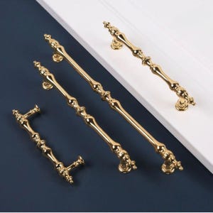 May include: Four gold cabinet pulls of different sizes. The pulls have an ornate, vintage design with decorative detailing. The hardware is displayed against a white and navy blue background, showcasing the polished finish and elegant style.