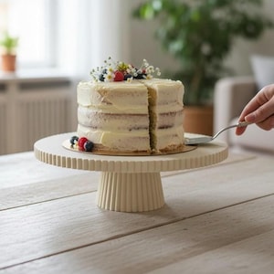 Könnte beinhalten: Ein runder, cremefarbener Tortenständer mit vertikal gerippter Textur am Fuß und am Rand der Platte. Der Ständer trägt eine mehrschichtige Torte mit weißer Glasur, die mit frischen Himbeeren, Blaubeeren und kleinen weißen Blüten verziert ist.