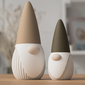 Könnte beinhalten: Zwei Zwergenfiguren mit texturierten Oberflächen. Der größere Zwerg hat eine beige spitze Mütze, während der kleinere eine olivgrüne spitze Mütze trägt. Beide haben gerippte weiße Körper, weiße strukturierte Bärte und kleine, glatte, helle Nasen.