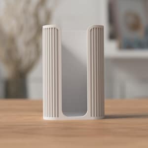 May include: A light beige, rectangular tissue box holder with vertical ribbed detailing on the sides. The holder is open at the top and bottom, and sits on a wooden surface. The background is blurred, with hints of a vase and a shelf.