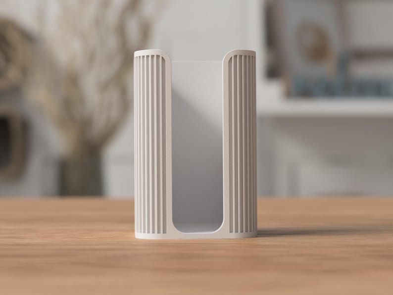 May include: A light beige, rectangular tissue box holder with vertical ribbed detailing on the sides. The holder is open at the top and bottom, and sits on a wooden surface. The background is blurred, with hints of a vase and a shelf.