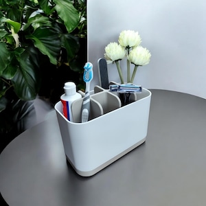 May include: A white and light grey rectangular bathroom organiser with multiple internal compartments. It holds a tube of toothpaste, a blue and white toothbrush, a dark grey nail file, a blue razor, and three white artificial flowers.