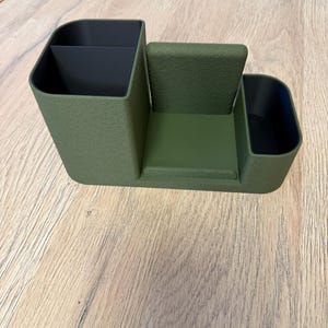 A textured, olive green desk organizer with black compartments and a central phone stand is viewed from the front on a light wood surface.