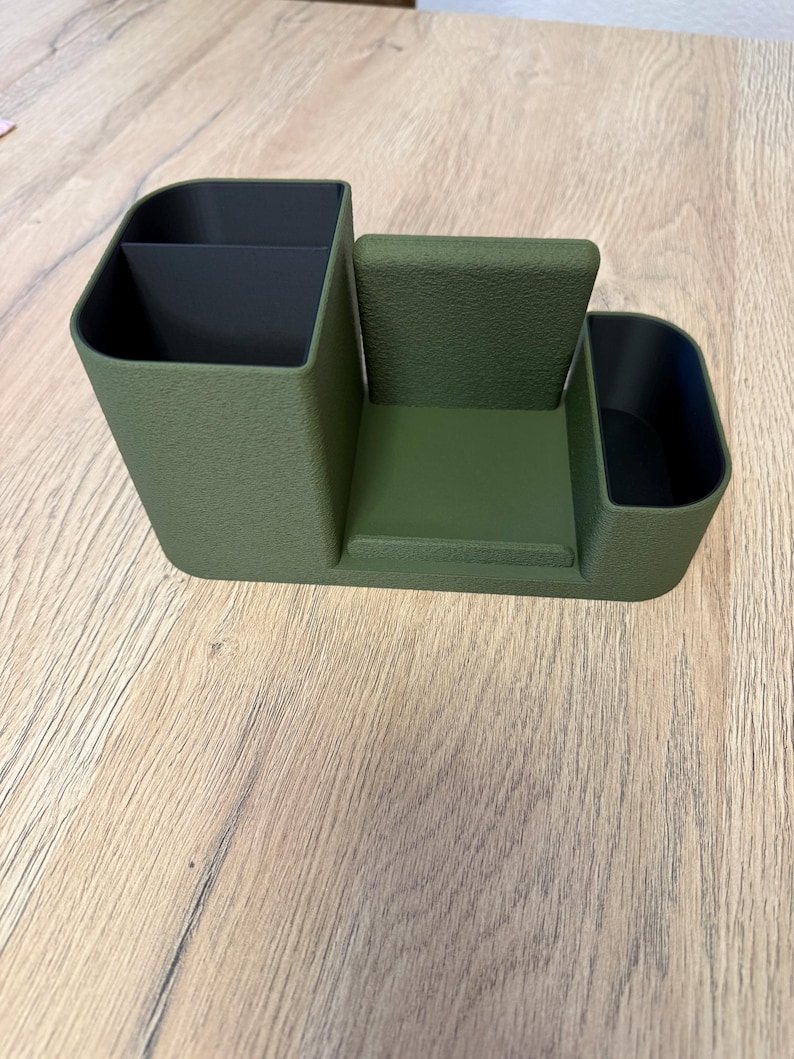A textured, olive green desk organizer with black compartments and a central phone stand is viewed from the front on a light wood surface.