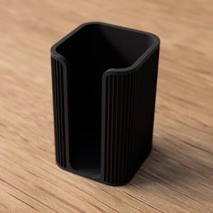 May include: A black, rectangular desk organizer with a vertical ribbed design. The open-top container is made of a solid material and sits on a wooden surface. The organizer is designed to hold items like pens or small office supplies.