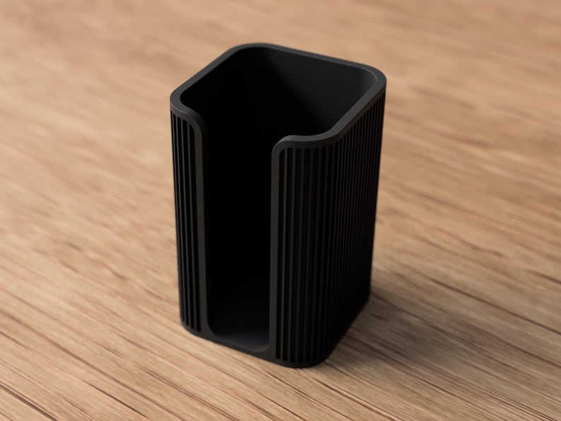 May include: A black, rectangular desk organizer with a vertical ribbed design. The open-top container is made of a solid material and sits on a wooden surface. The organizer is designed to hold items like pens or small office supplies.