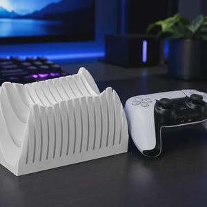 Könnte beinhalten: Ein weißer Controller-Ständer aus Kunststoff mit einer Reihe vertikaler, paralleler Lamellen zur Aufnahme eines Gaming-Peripheriegeräts. Neben dem Ständer befindet sich ein weiß-schwarzer kabelloser Gamecontroller mit zwei Analogsticks.