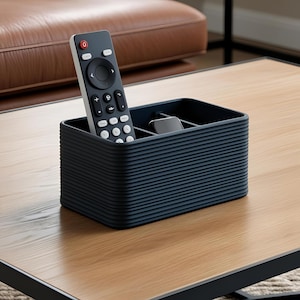 Puede incluir: Un organizador de almacenamiento rectangular de color gris oscuro con una textura exterior acanalada y divisores internos. El organizador contiene un control remoto negro con botones blancos y un pequeño dispositivo electrónico negro con pantalla.