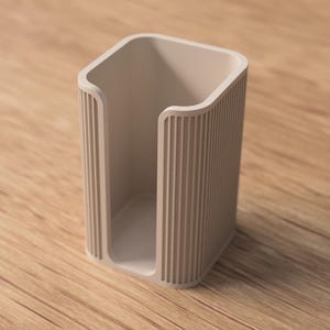May include: A beige, rectangular holder with vertical ribbed detailing on the sides. The open-top container is set on a wooden surface. The holder's design is minimalist and modern, suitable for various uses.