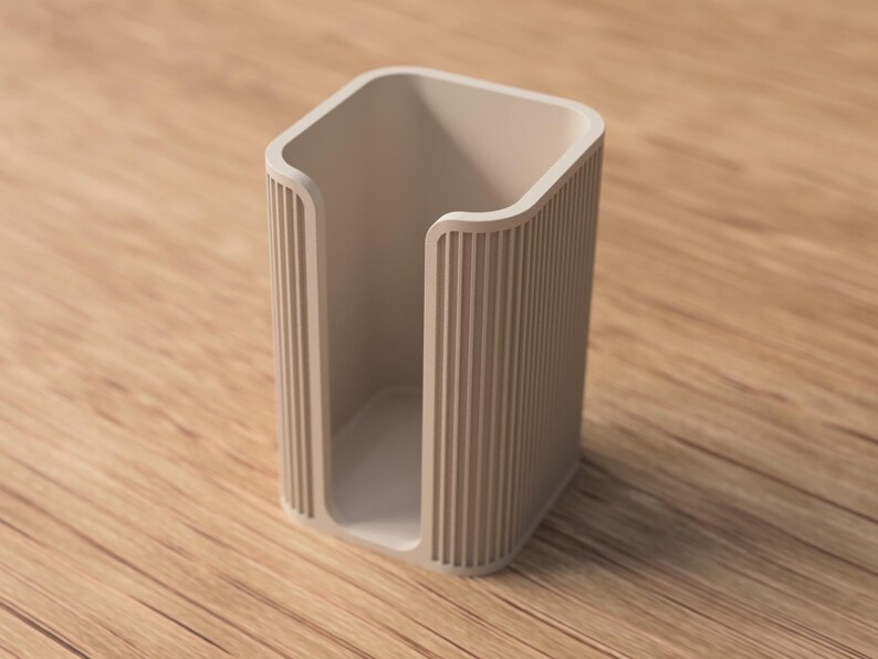 May include: A beige, rectangular holder with vertical ribbed detailing on the sides. The open-top container is set on a wooden surface. The holder's design is minimalist and modern, suitable for various uses.