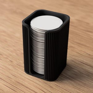 May include: A black, rectangular coffee filter holder with a stack of white, circular filters inside. The holder has vertical grooves on the sides and sits on a wooden surface. The design is modern and minimalist.