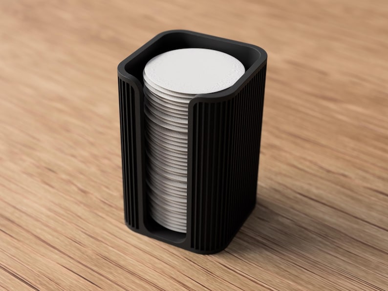 May include: A black, rectangular coffee filter holder with a stack of white, circular filters inside. The holder has vertical grooves on the sides and sits on a wooden surface. The design is modern and minimalist.