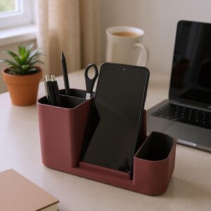 A dark pink desk organizer with a smartphone, pens, and scissors is on a wooden desk with a laptop, coffee, and plant.