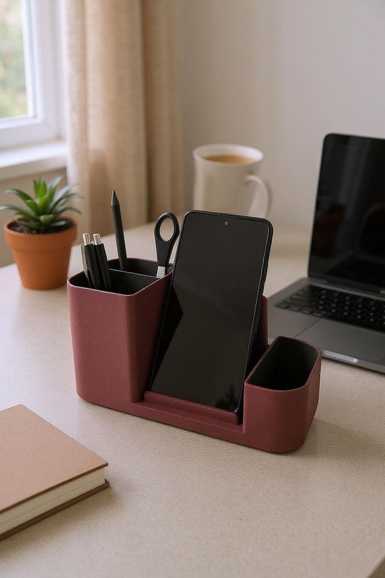 A dark pink desk organizer with a smartphone, pens, and scissors is on a wooden desk with a laptop, coffee, and plant.