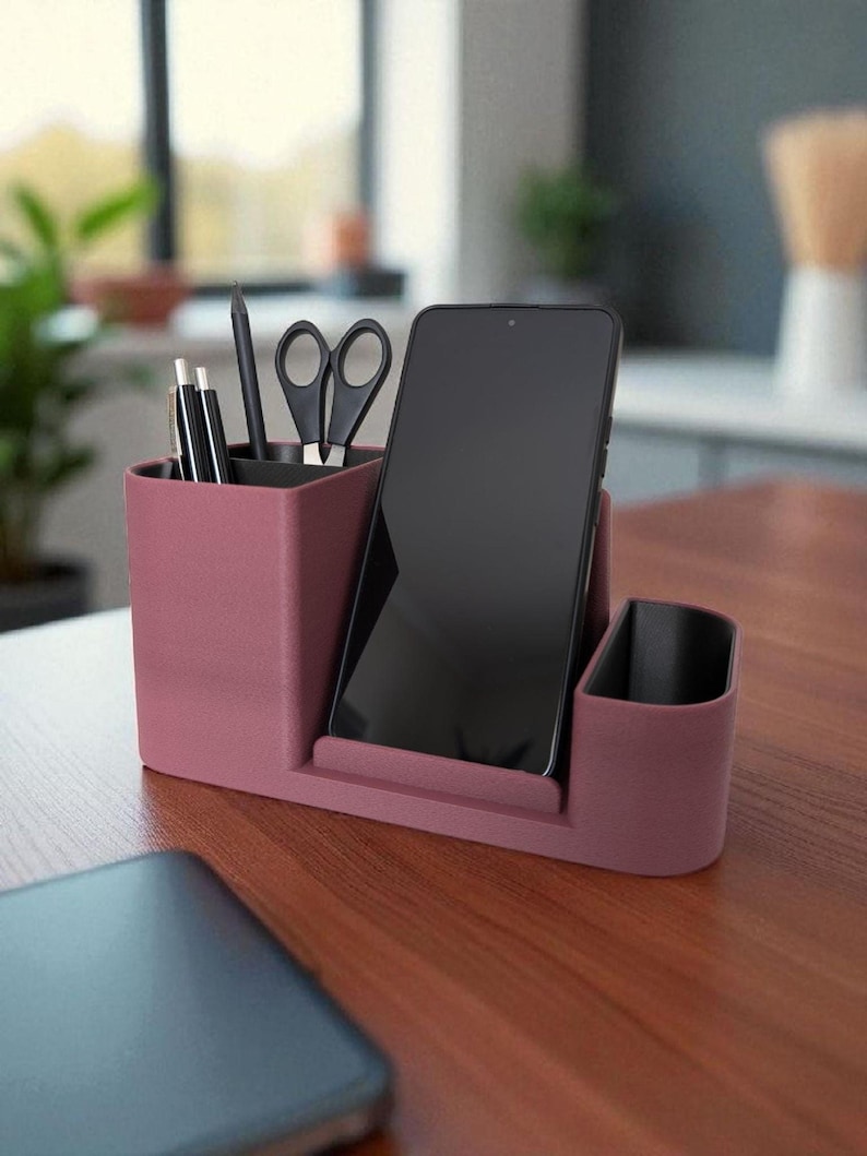 A textured, dark rose-colored desk organizer with a smartphone, black pen, and scissors in its compartments, on a wooden desk.