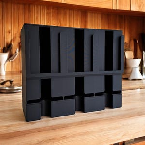 May include: A black, multi-compartment organizer for kitchen or office use. The organizer has multiple vertical slots and lower compartments, designed to hold various items. It is made of a matte black material and sits on a wooden surface.