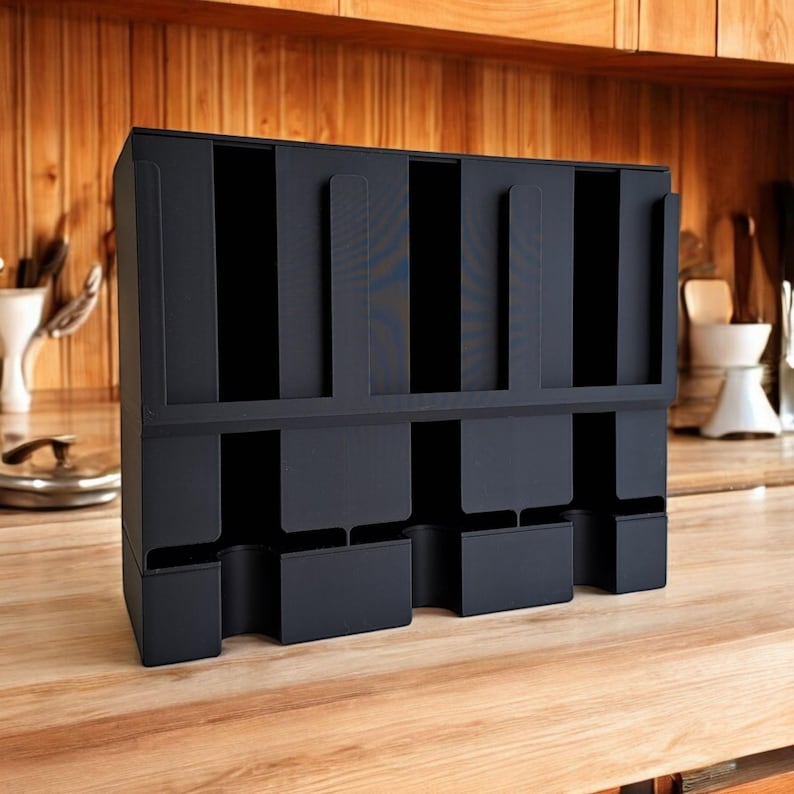 May include: A black, multi-compartment organizer for kitchen or office use. The organizer has multiple vertical slots and lower compartments, designed to hold various items. It is made of a matte black material and sits on a wooden surface.