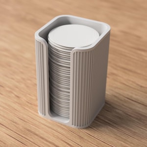 May include: A light gray, rectangular holder with vertical ribbed sides, containing a stack of white, circular objects. The holder has a curved top and is set on a wooden surface. The design is minimalist and modern.