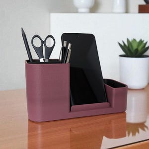 A textured, dark rose-colored desk organizer with a smartphone, black pen, and scissors in its compartments, on a wooden desk.