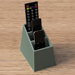 Puede incluir: Un soporte para mandos a distancia de color verde salvia con una textura exterior acanalada y tres compartimentos verticales. El soporte contiene tres mandos negros de distintas alturas, cada uno con varios botones y forma rectangular.