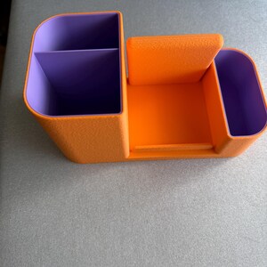 An orange desk organizer with purple compartments and a central phone stand is viewed from the front on a gray surface.