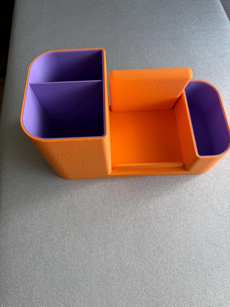 An orange desk organizer with purple compartments and a central phone stand is viewed from the front on a gray surface.