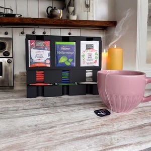 May include: A black tea organizer with three tea packet displays, each holding different tea flavors. A pink teacup sits on a wooden surface, with a tea bag and two lit candles in the background. The tea packets are labeled with German text.