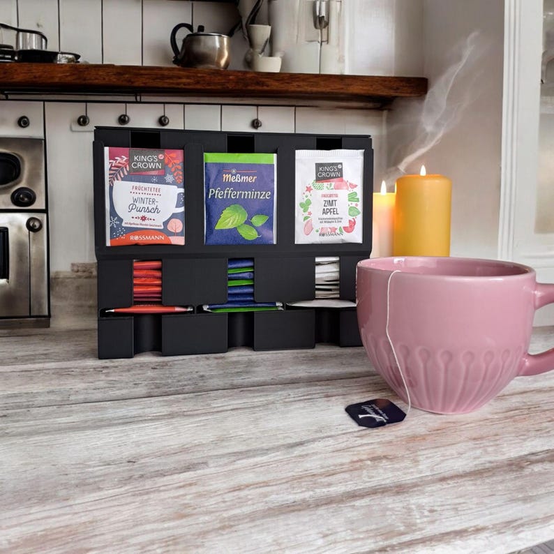 May include: A black tea organizer with three tea packet displays, each holding different tea flavors. A pink teacup sits on a wooden surface, with a tea bag and two lit candles in the background. The tea packets are labeled with German text.