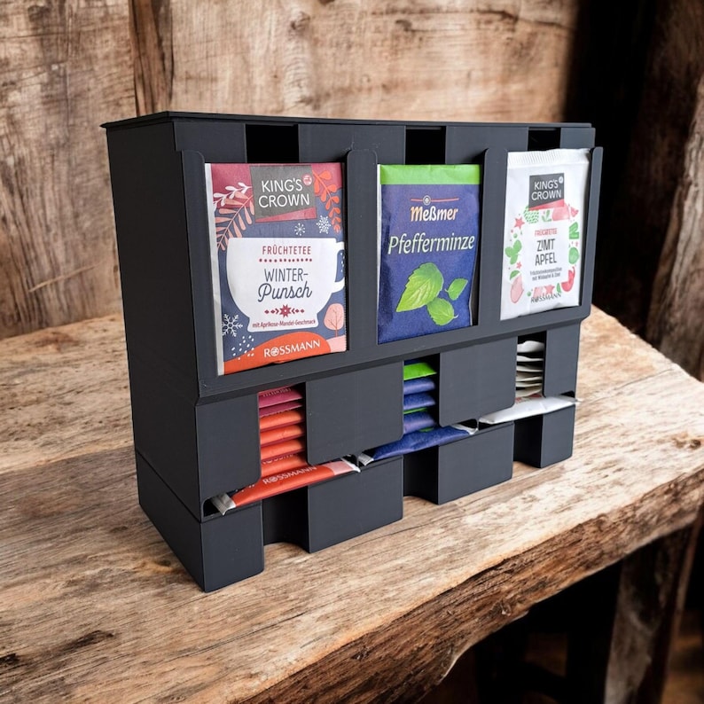 May include: A black tea bag organizer with three compartments for tea bags and three slots for tea packets. The organizer has the text "KING'S CROWN" and "ROSSMANN" on the tea packets. The tea packets are in various colors.