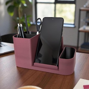 A dark pink desk organizer with a smartphone, pens, and scissors sits on a wooden desk next to a coffee cup and laptop.