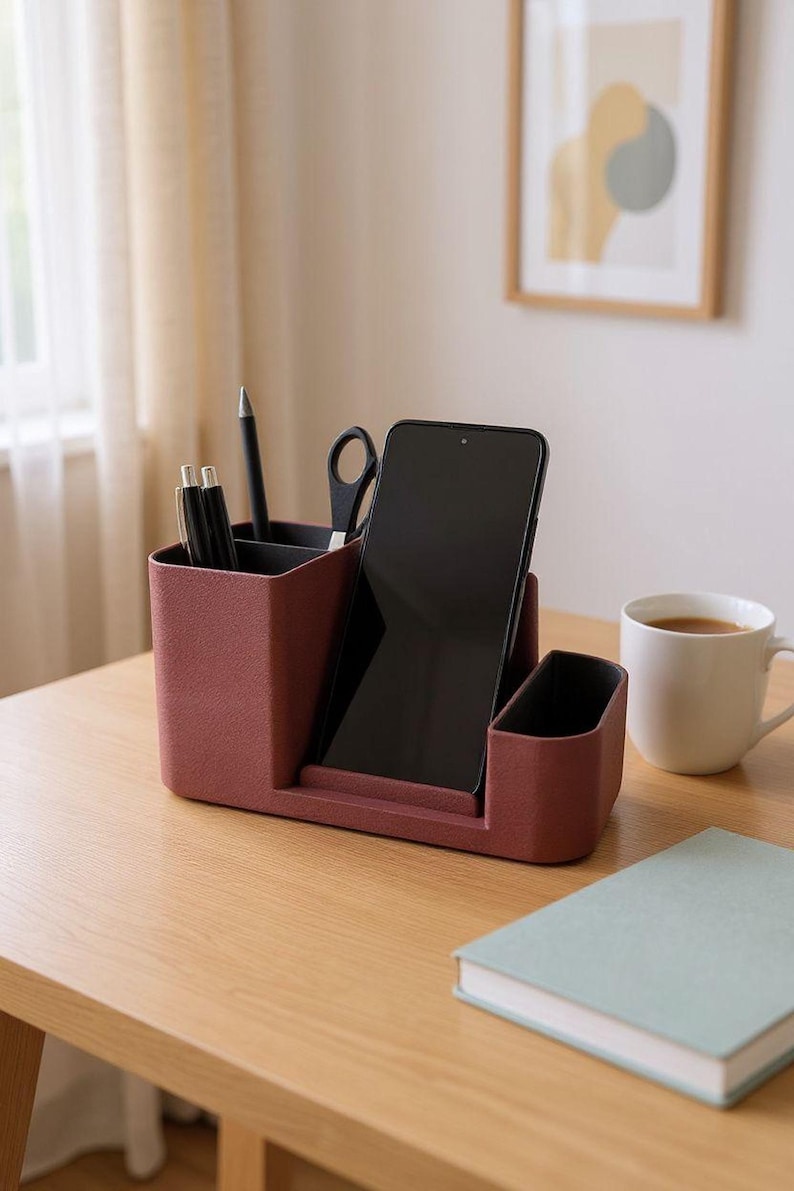 A dark pink desk organizer with a smartphone, pens, and scissors is on a wooden desk with a coffee cup and a light blue book.