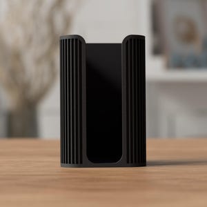 May include: A black, rectangular holder with vertical ribbed detailing on the sides. The holder is made of a matte material and sits on a light brown wooden surface. The background is blurred, suggesting an indoor setting.